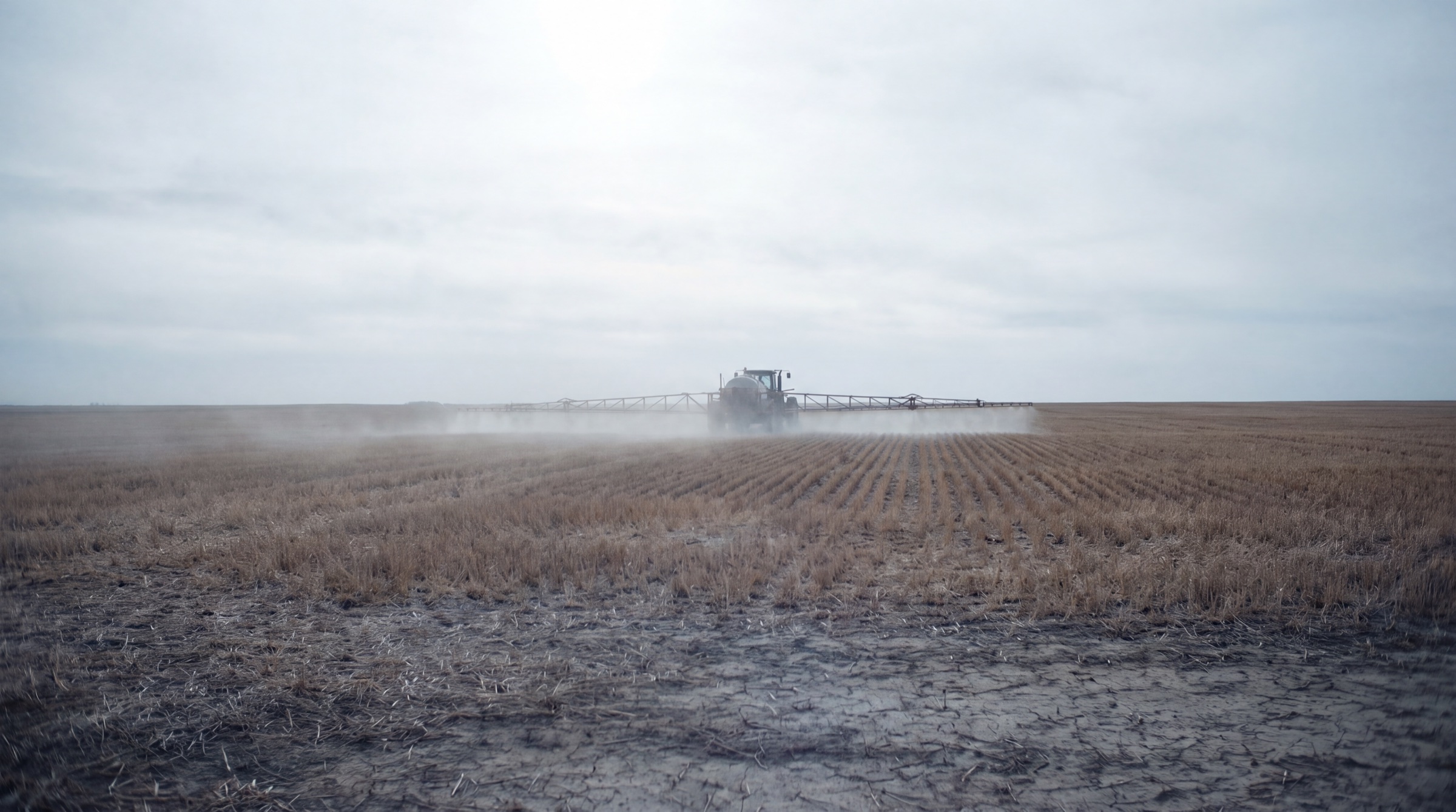 A crop sprayer working a monoculture field, chemical mist drifting across bare soil